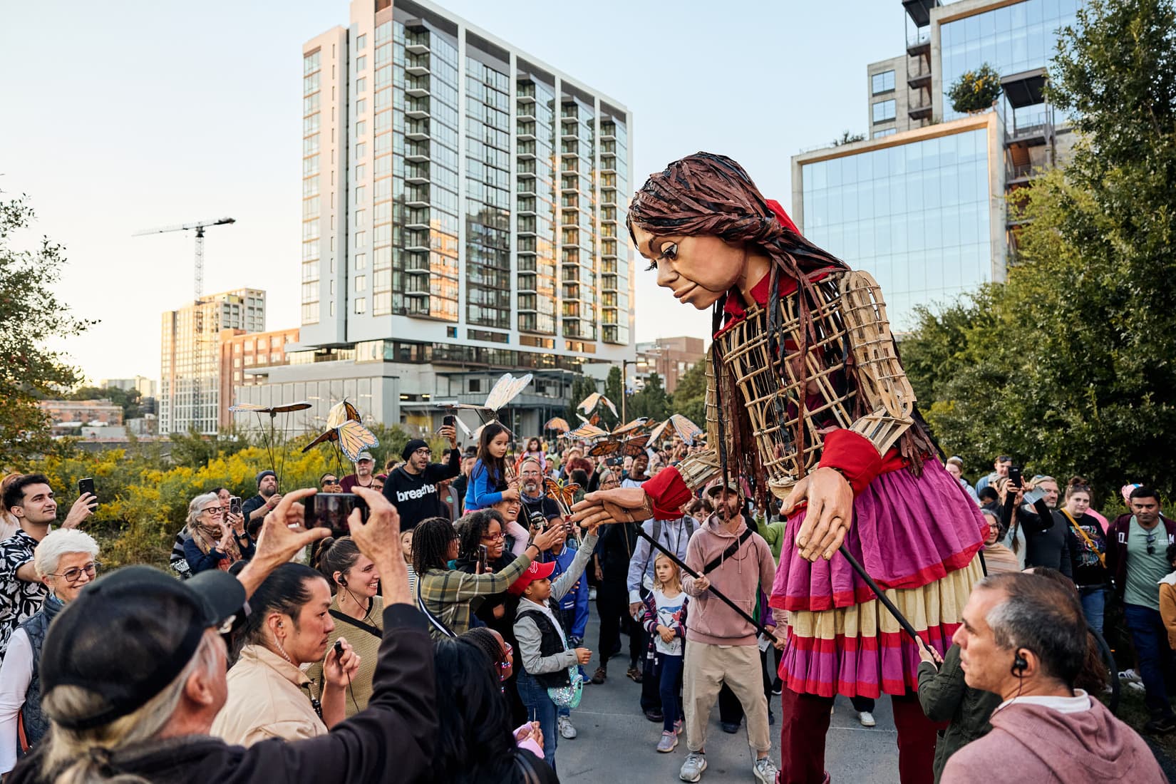 A 12-foot wooden puppet depicting a young girl stands on the Eastside Trail, surrounded by a crowd of people smiling and taking photos.