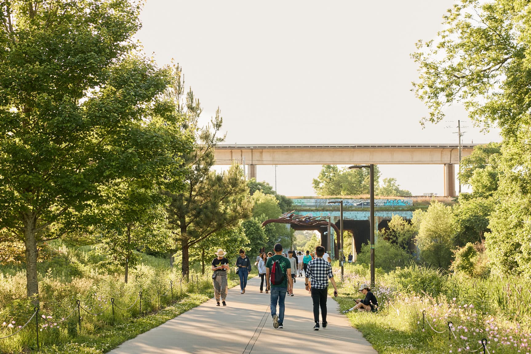 People walk down a paved pathway, surrounded by greenery on either side.