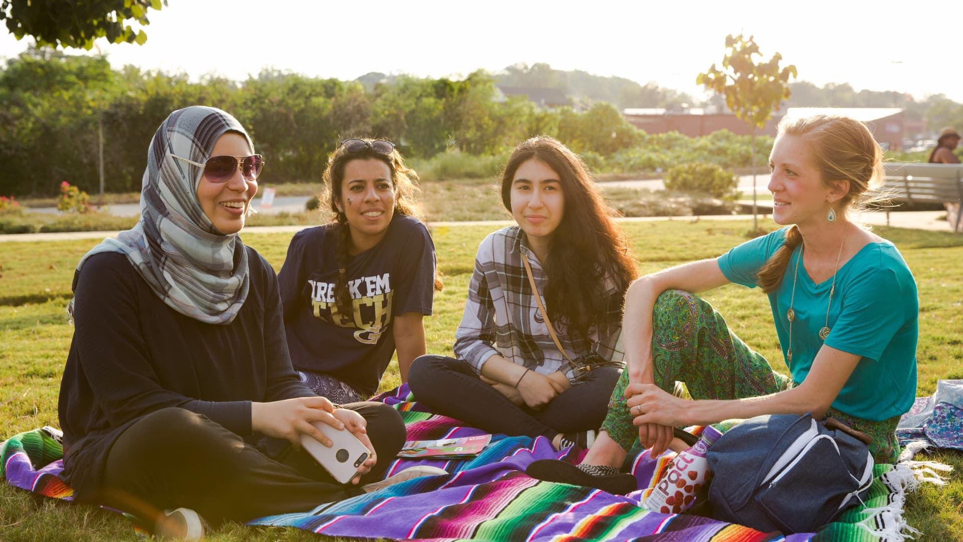 Four people sit on a picnic blanket, talking and laughing.