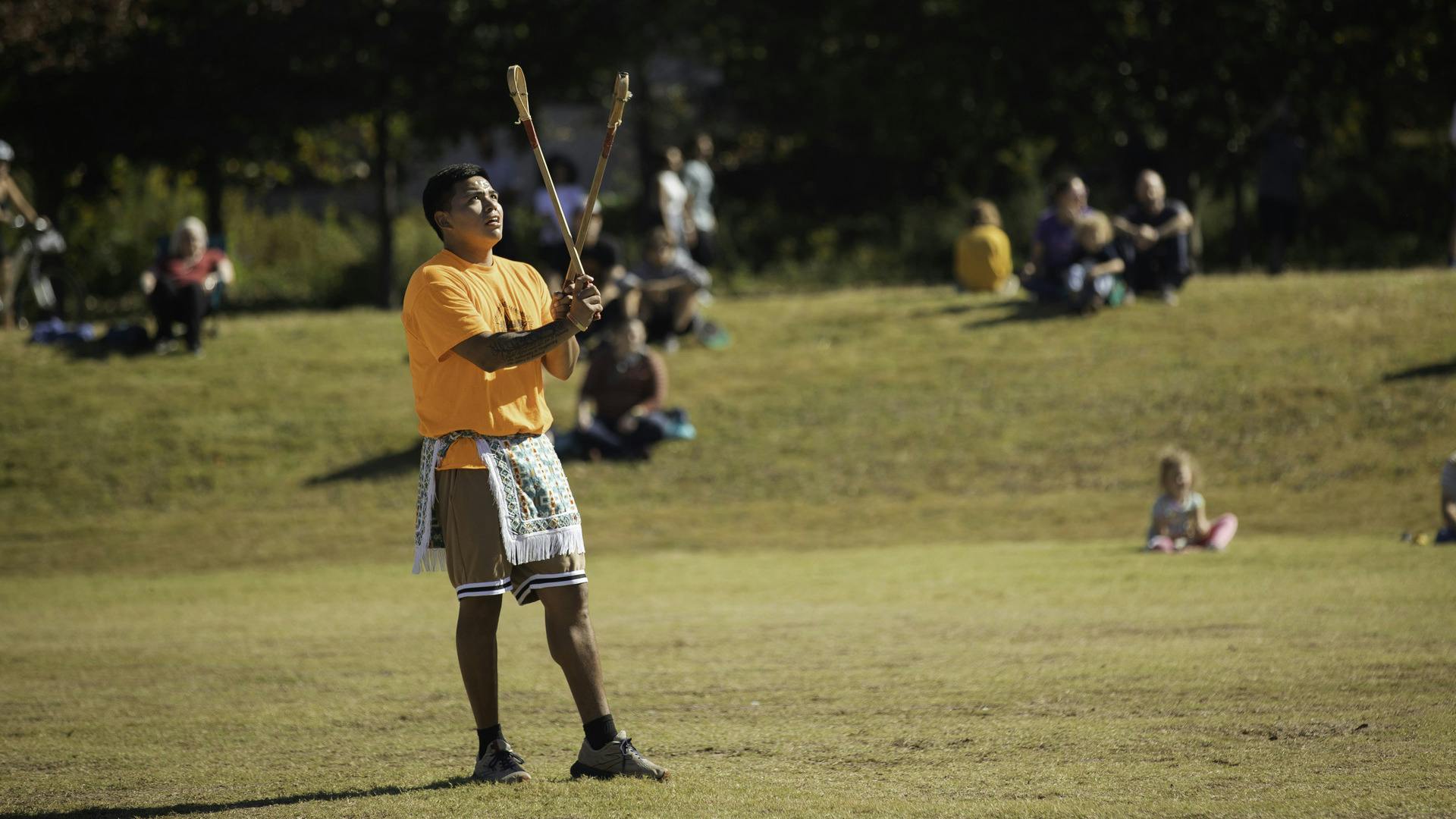 A person stands, holding two toli sticks, with a ball in the air. Nearby, people watch while sitting on a hill.